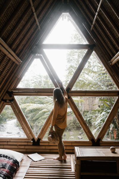 Woman stands inside a cozy bamboo house, gazing at lush greenery through the window.