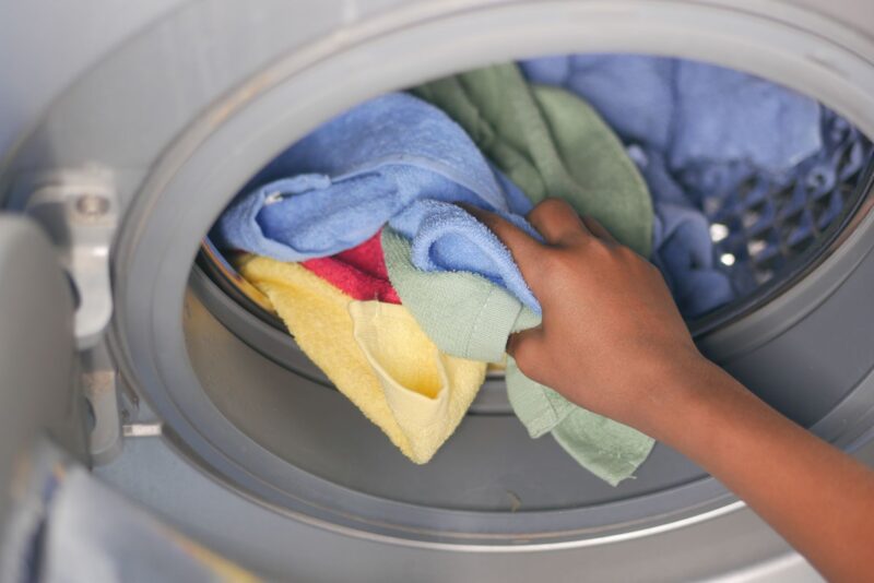 A close-up of a hand loading="lazy" colorful towels into a front-loading washing machine.