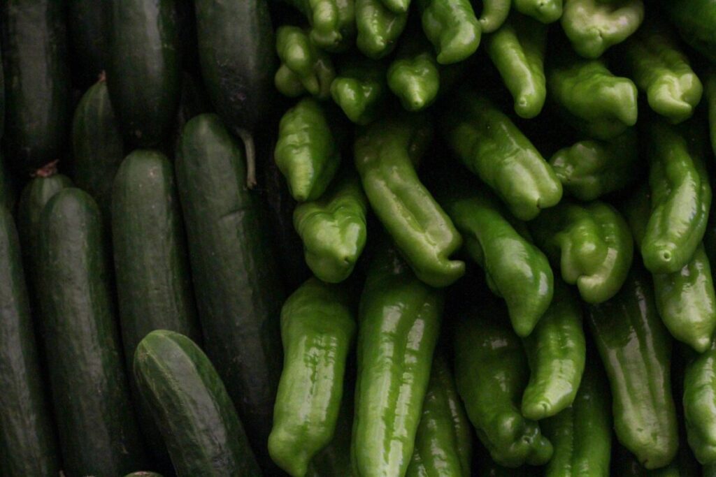 A close-up shot of fresh green cucumbers and Anaheim peppers at a market display, showcasing vibrant green hues.