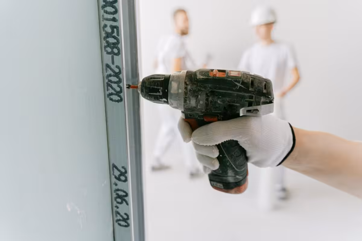 A construction worker uses a drill on drywall with blurred workers in the background.