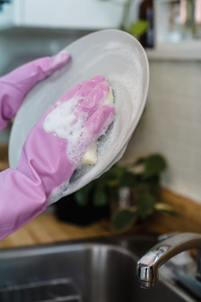 Hands in pink gloves washing a plate with a sponge in a kitchen sink.